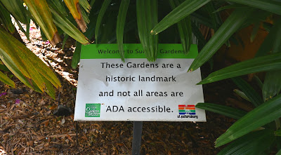 A sign, located amongst some ferns and before the entrance to Sunken Gardens, that reads, "Welcome to Sunken Gardens. These Gardens are a historic landmark and not all areas are ADA accessible.