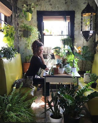 A woman seated on a yellow bench-type seat at a table surrounded by plants.