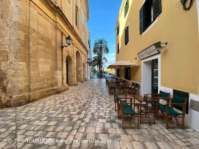 Rows and rows of wooden and dark green director chairs at wooden bistro tables along the wall of a pastel yellow period property with green wooden shutters and a white awning above its entrance and the words Bar Imperi, opposite the entrance of a cathedral, in a lane that leads to a large square lined by giant palm trees under a blue sky.