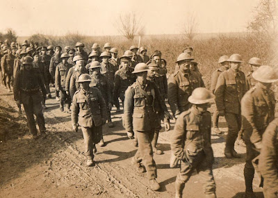 A sepia photograph of dozens of British soldiers, identifyable by their tin hats, trudging unarmed down a dusty road, guarded by German soldiers