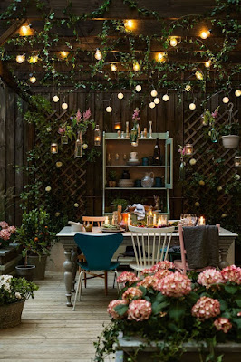 A wooden table with mismatched chairs in an arbor full of flowers and twinkly lights.