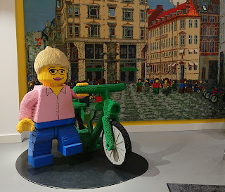 Large scale model of person with bicycle, with a picture of a city square in the background, all made from LEGO.