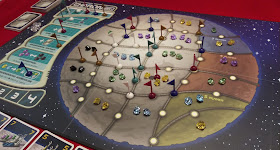 A view of the board in the middle of a game. Gems of various colours have been placed in the spaces on the board, and a number of plastic flags in various colours have been placed on the icons on the borders between the spaces. One flag in each colour remains on the special actions spaces, and one flag of each colour occupies the spaces of the player order track in the top left.