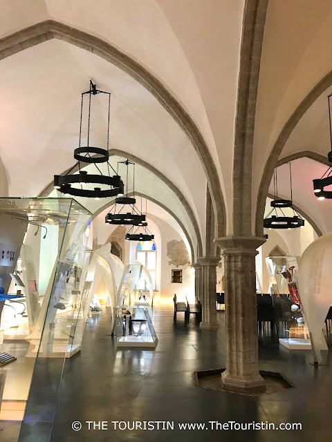 Five large glass exhibition stands in a large room under a white vaulted stone ceiling decorated with medieval-style ceiling lights.