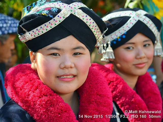 Matt Hahnewald; Facing the World; Asia; Northern Thailand; Lu-mien; Yao; travel destination; tourism; hilltribe; ethnic minority; ethnic; traditional Yao costume; traditional Yao headdress; Doi Hua Mae Kham; festival; girl; world cultures; red pompoms; Golden Triangle; smiling; eye contact; street portrait; closeup; double portrait; Nikon DSLR D3100; 50 mm prime lens