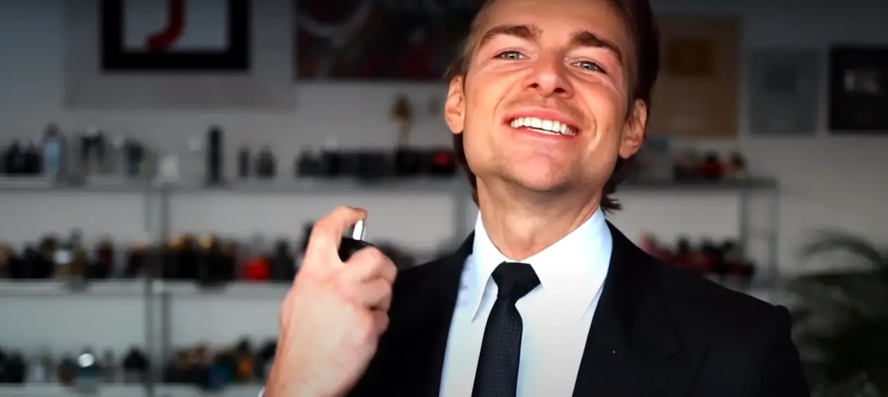 A man in a tailored black suit and coat sprays cologne onto his neck. The mist catches the light, creating a delicate, fragrant cloud around him. His poised and elegant appearance, combined with the act of applying cologne, exudes sophistication and confidence. The scene captures a moment of refined personal grooming.