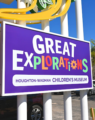 A purple sign, with white and multi-colored text that reads, "Great Explorations." Below that, with a white outline, is purple text that reads, "Houghton-Wagman Children's Museum"