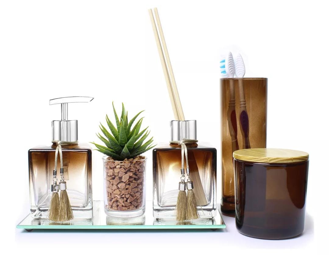 Fundo branco. Conjunto de banheiro: sobre uma bandeja espelhada, tem um dispenser para sabonete líquido, um vaso com pedrinhas marrons e planta tipo suculenta verde, e um frasco difusor com palitinhos. O dispenser e o frasco são em degradê marrom e transparente, de tampa prata, com cordões bege em volta das tampas, com detalhes prateados e pompom de franja bege na ponta. Ao lado, tem um copo marrom translúcido com duas escovas de dente e um porta-algodão da mesma cor do copo, com tampa bege.