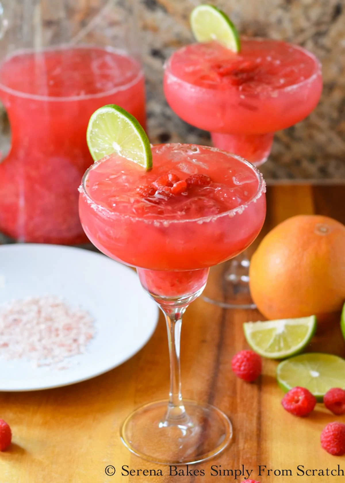 A side shot of 2 Raspberry Margaritas in tall Margarita Glasses with a glass pitcher of Raspberry Margaritas to the left hand side and a small white plate of pink salt on a wooden cutting board.