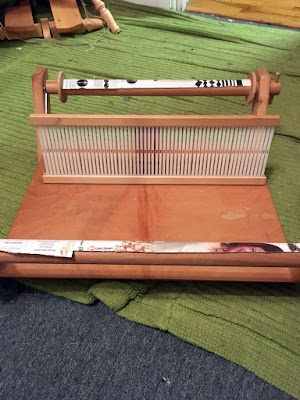 A small wooden rigid heddle loom, with pieces of magazine paper wrapped over the front and back beams, sitting on a green rug.
