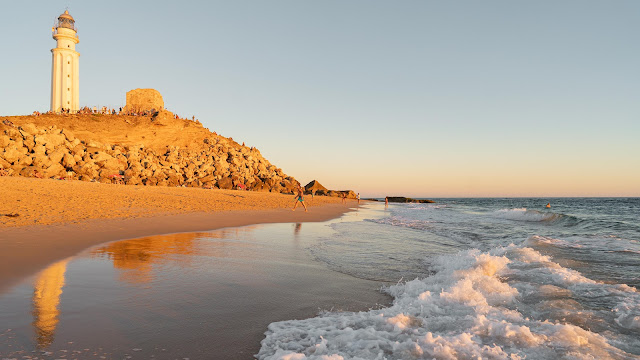 Orilla de playa con olas doradas por los rayos de sol al atardecer, el reflejo de un faro en el agua mojada y un pequeño acantilado con un faro.