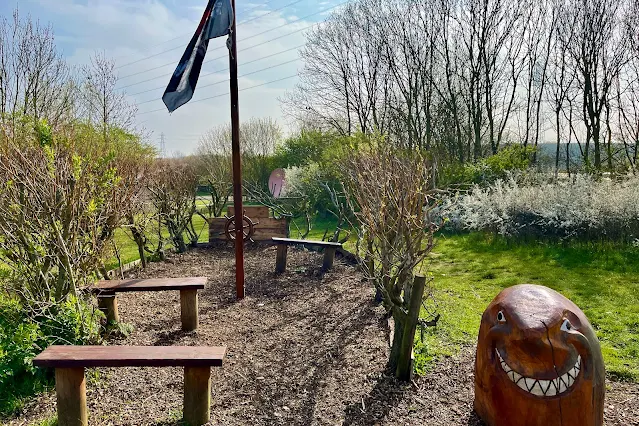 some benches, surrounded by plants in a boat shape, a flag and a ships wheel