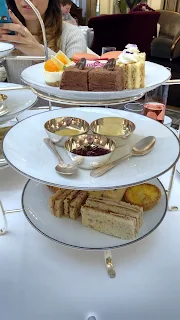 3 tiered stand with a white plate on each tier. The bottom tier has small sandwiches with the crust cut off. The center tier has stainless steel containers of jams. The top plate has an assortment of cakes.