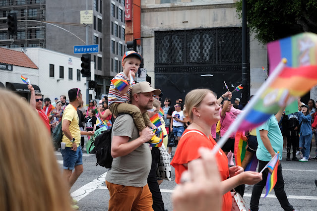 Pride paradegoers walking down the street.  A small child is carried by an adult man (father?) while he chews on a small pride flag.