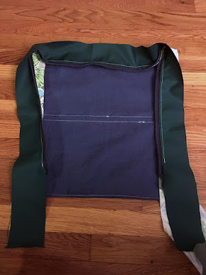 The back half of a backpack, showing white stitching horizontally across the blue lining where the pieces were joined, and whit a dark hunter green zipper panel lined in green, teal, and white tropical floral fabric, laid flat on a wood floor.