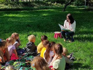 Grupa dzieci siedzi na kocu, który rozłożony jest na trawie. Dzieci słuchają pani bibliotekarki, która czyta im bajkę.