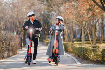 Source: Neuron Mobility. A man and a woman look at each other while posing on Neuron Mobility e-scooters in an autumn scene., Korea.