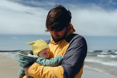 समुद्र के किनारे एक व्यक्ति खड़ा हुआ है जिसने अपनी गोदी में एक छोटे बच्चे को लिया हुआ है