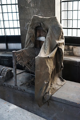 A battered chair, its cane seat unravelling, draped with a worn, pale brown coat.
