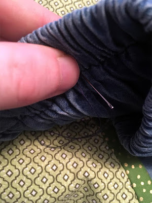 A close-up of a needle inserted through a blue denim skirt just below the waistband and into a green print pocket.