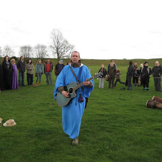 Bard wearing a blue robe in circle at Avebury stone circle with people around him