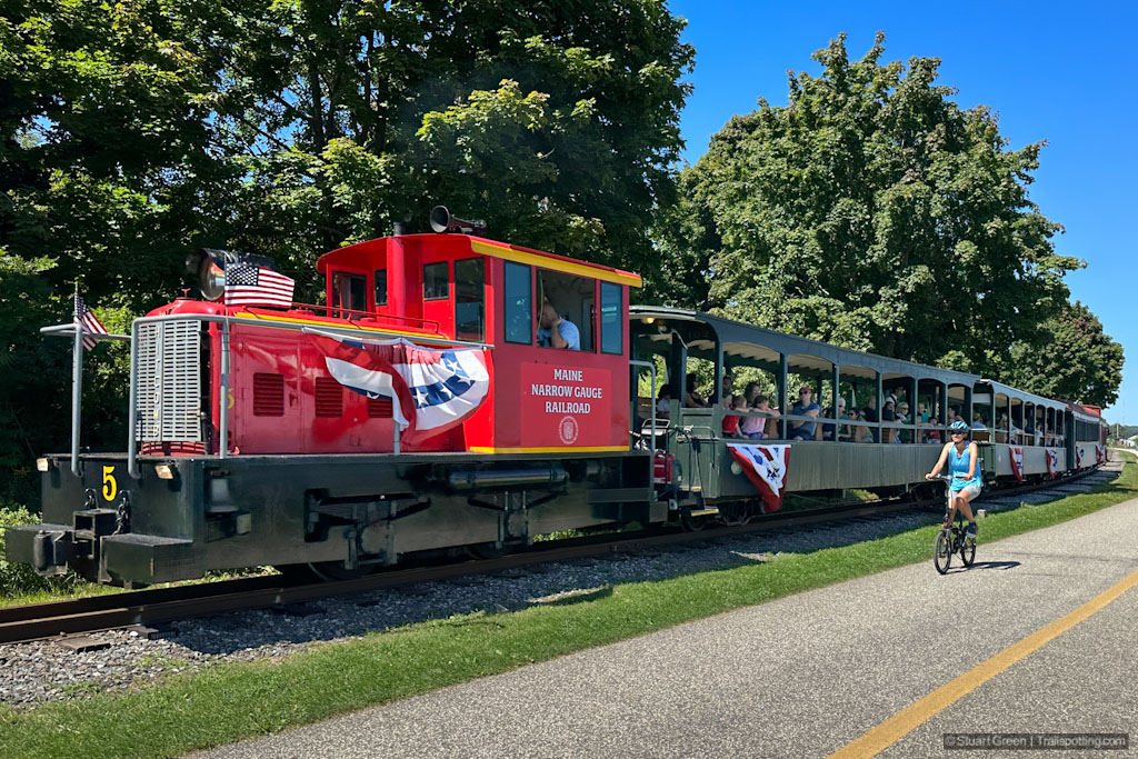 Cyclist riding on a rail trail adjacent to a narrow gauge railroad train.