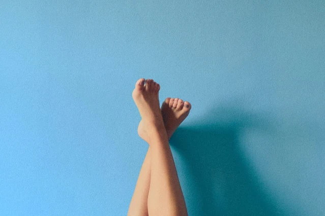 A woman's legs and feet emerge from the bottom center and rest crossed in the center of this photograph against a blue background.
