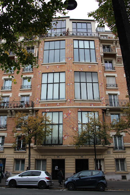 Photo of a five-story building. The ground floor is plain sandstone, the upper stories are decorated brickwork. Each floor above the ground floor has two very large windows right in the center, reaching, very likely, from the floor to the ceiling. These are multipaned, handsome windows. Each floor might have an artist's studio on it. The other windows are normal size - maybe there are more than five floors to the left and right of the center section with the big windows.