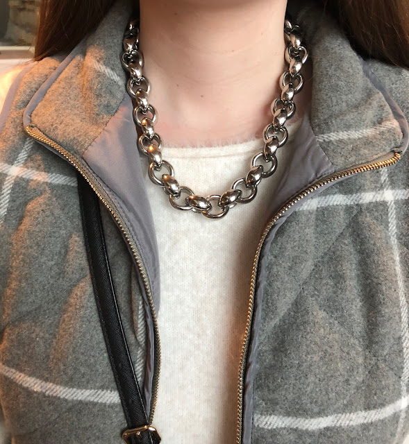 Model is wearing grey vest with white sweater and black and silver accessories.