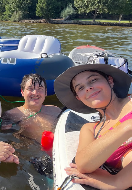 A selfie of you and Andy. You have dad's fisherman hat on with your orange bikini top. You are laying down on your paddle board. Andy is in the water swimming next to you. The floats are all hooked together.