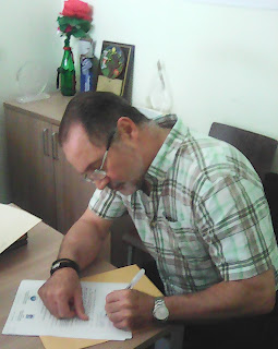 En la foto Mario Lebrón sentado, firma el formulario de autorización. Mario viste una camisa de mangas cortas a rayas negras, marrones, verde clara y oscuro. Su pelo es color negro con entradas pronunciadas, cara redonda, con gafas recetadas. Al fondo sobre una credensa, algunos reconocimientos recibidos por USEPEDI y una flor de papel en una botella