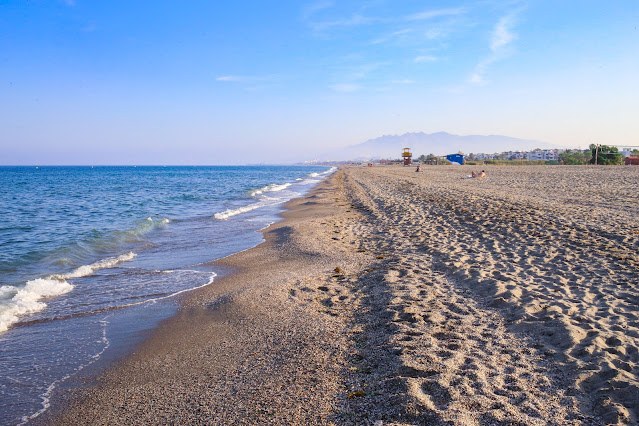 Playa de arena fina amplia y llana con las azules aguas del mar a su frente