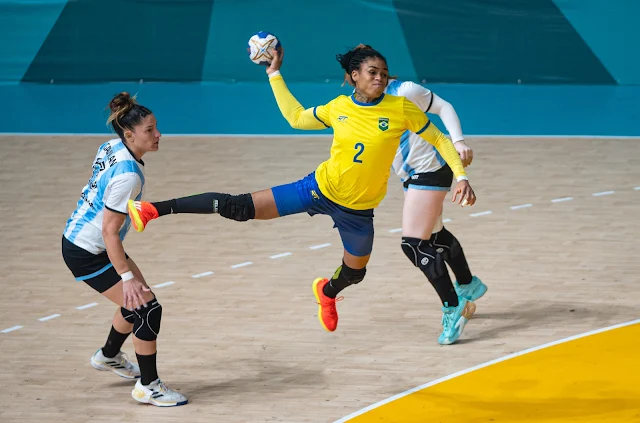 Jogadora do Brasil com a número 2, salta para arremessar a bola para o gol. Ela é negra e está com a camisa amarela entre duas argentinas