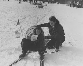 A black and white photograph of a man crouched by a girl on skis.