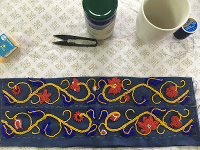 A pair of rectangles of blue linen with similar but unmatched mirrored vining designs in yellow, blue, red, white, and black beads, laid out on a white tablecloth with small thread snips, a jar of blue beads, and a tea mug nearby.