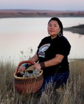 A brunette woman standing in a field with a large basket full of plants. There is a lake in the background, Linda Black Elk