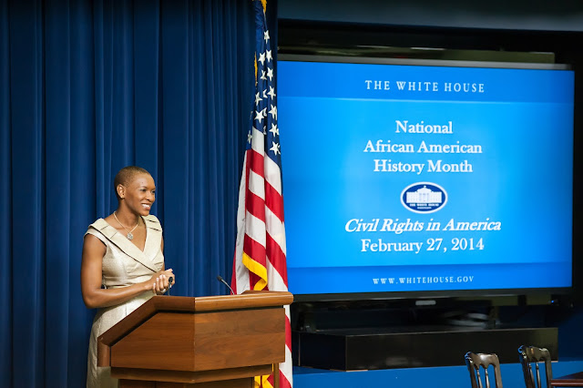 Claudia Gordon frente a un atril, bandera de USA y una proyección a un lado sobre la National African American History Month