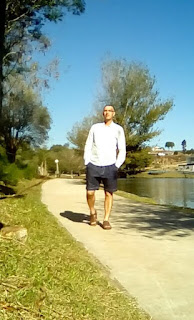 Foto pessoa de frente, numa pista de caminhada ao ar livre, com sandália marrom, bermuda azul marinho e camisa xadrez clara!