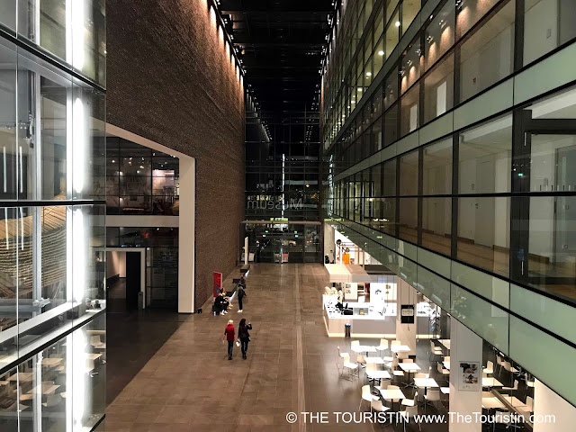 Six people sit and two walk through a massive hallway with exposed red brick and four floors with huge glass windows on both sides.