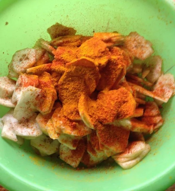 Close view of sliced green bananas, chili powder, turmeric powder and salt in a green plastic container.