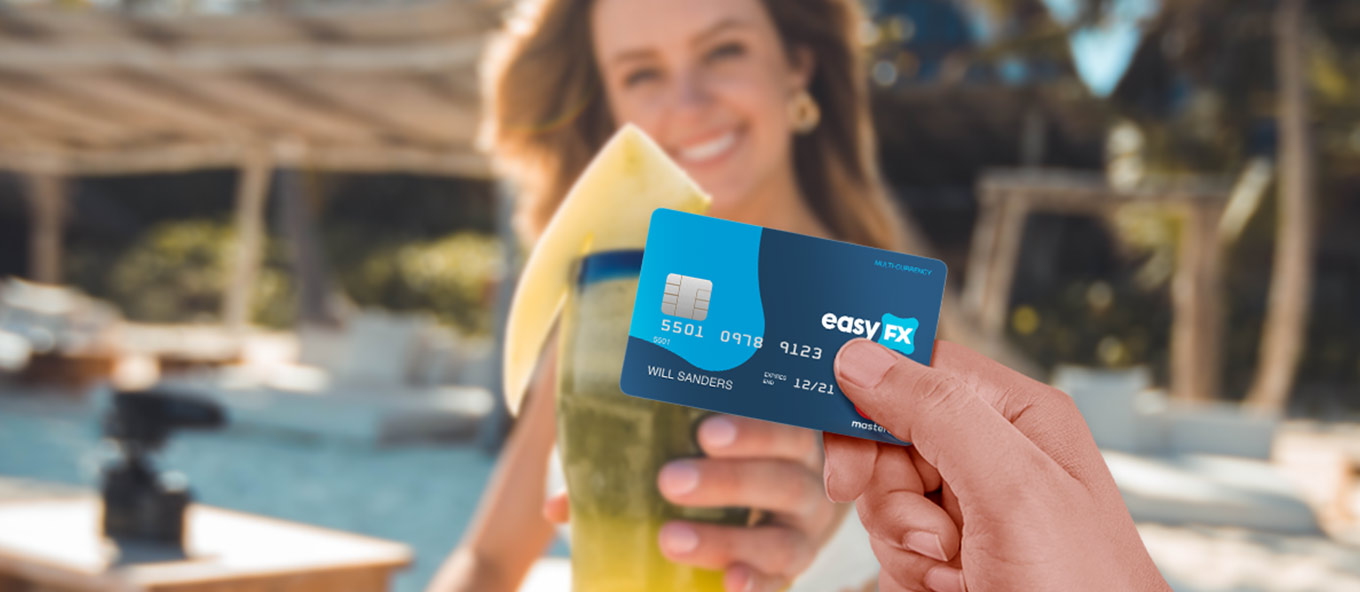 A POV of a person holding an easyfx travel money card infront of another person, smiling and holding a drink in a holiday setting in the sun.