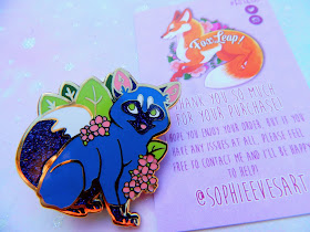 A photo of a enamel pin by FoxLeap! of a blue fox with flowers and leaves surrouding it. it has a glittery tail and paws.