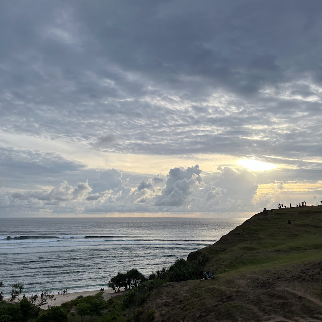 Pemandangan matahari terbenam di pinggir laut dengan bayangan-bayangan orang di atas bukit