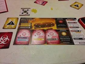 The yellow player's Evolution Slide. An image of a magnified bacterium on the top, with the 'core traits' (two listings of 'Infectivity +1' and one listing of 'Lethality +1') in the top left corner, five spaces to place Trait Cards (two of which, currently unoccupied, grant abilities so long as they remain unoccupied), three of which contain Trait Cards, and a Turn Summary listing along the right edge of the mat. The trait cards have a cost/point value in the upper left corner, next to which is the card's name (these three are Confusion, Mass Hysteria, and Blindness), and one or more abilities listed on the bottom (Confusion has 'Airborne' and 'Cold Resistance,' Mass Hysteria has 'Infectivity +1,' and Blindness has 'Lethality +1'). Some Event cards, unused Trait Cards, and a claimed Country Card, as well as some of the hexagonal yellow tokens, can be seen on the table nearby.