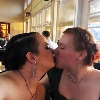 Two young women in evening dresses, smiling through a kiss, in a light-filled ballroom.