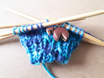 Recently-started knitting project on double-pointed needles with a stitch marker made of a miniature cake with a slice taken out of it.