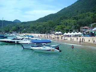 Playa de arenas firmes y distintos bares con techos de paja, en el agua calma de mar varias lanchas ancladas cerca de la costa.