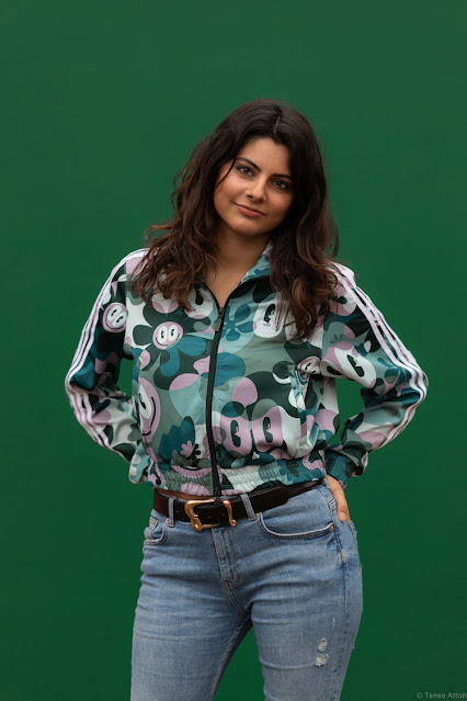 Mulher mestiça, de origem italiana e paquistanesa, vestindo calças jeans e blusa estampada com tons de verde. Utiliza um cinto de couro com fivela dourada. Está posicionada de frente a um fundo totalmente verde.