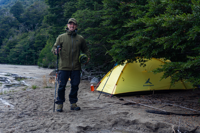The picture shows a man with hiking sticks on the beach in front of a yellow tent.