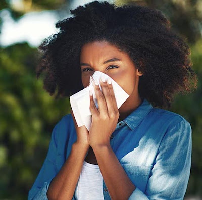 Image: a Black woman sneezes into a tissue due to outdoor allergies